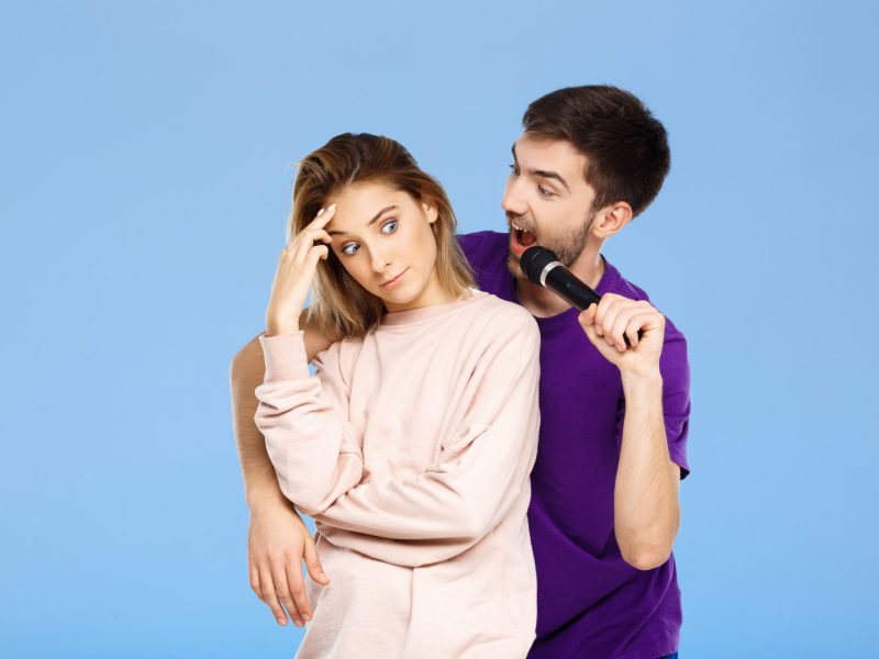 Young beautiful couple over blue background. Man singing in microphone girl didpleased. Copy space.